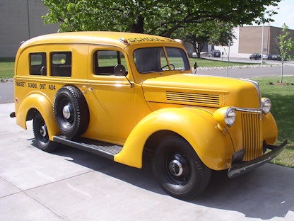 A yellow collector school bus parked in a driveway, with a tree and buildings in the background.