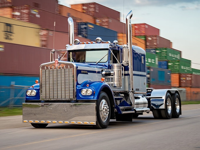 Blue collector commercial truck on a road, with shipping containers in the background.