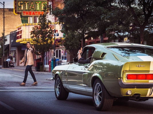 A person in a well-aged collector sports car and a woman crossing a downtown street give each other a thumbs up.