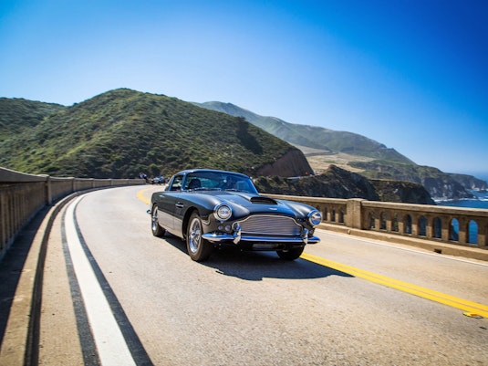 A well-aged collector car cruises down a scenic highway next to a large body of water. Behind the car are green, rolling hills and a blue sky.