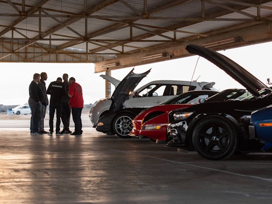 A group of men converse together under a large parking canopy, next to an angled row of parked collector cars with hoods up.