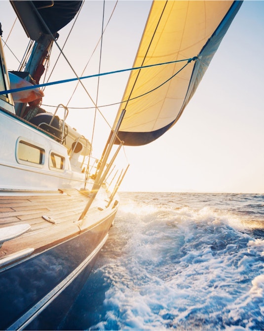 A yacht is directed through water with a clear and sunny sky backdrop.