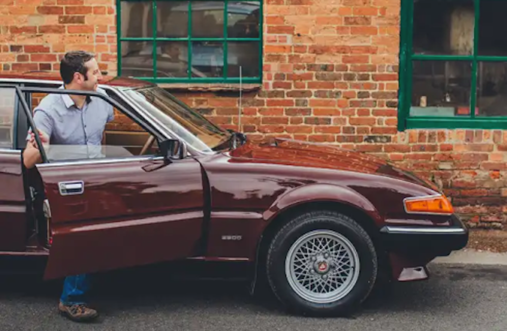 A collector car with the driver's side door open with a man getting into the vehicle next to a brick building